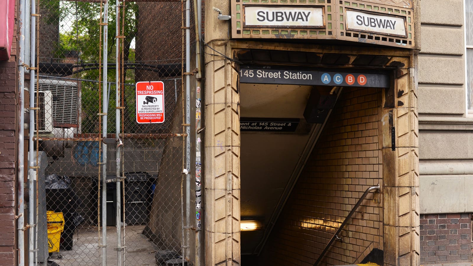 Photograph of a subway entrance set into a stone building, with two old Subway signs above, a sign reading 145 Street Station with A, C, B and D line symbols, tiled stairs going down, and a fenced alley with a No Trespassing sign beside it