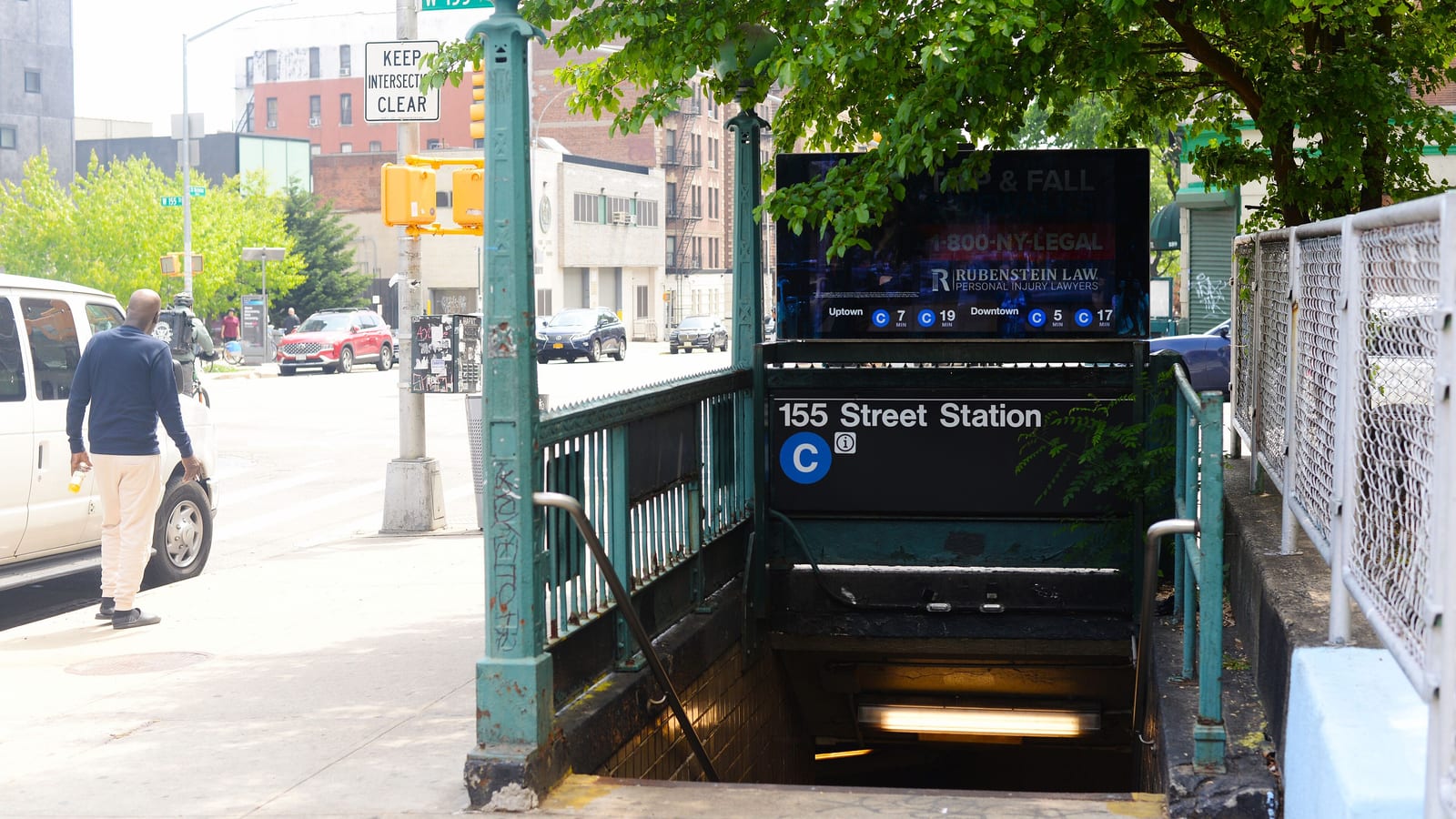 Photograph of a green iron subway stair entrance with a sign reading 155 Street Station and a blue C line symbol, a digital screen above showing train times, a tree overhead, a man walking away along the sidewalk at left and cars on the avenue behind