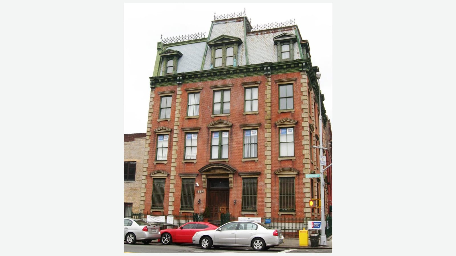 Photograph of a four-story red brick building with stone corner blocks, arched stone window heads, a slate mansard roof with three dormers and iron cresting, the number 1854 beside a wooden double door, an iron fence and three parked cars, on a cloudy day, shown on a plain light background