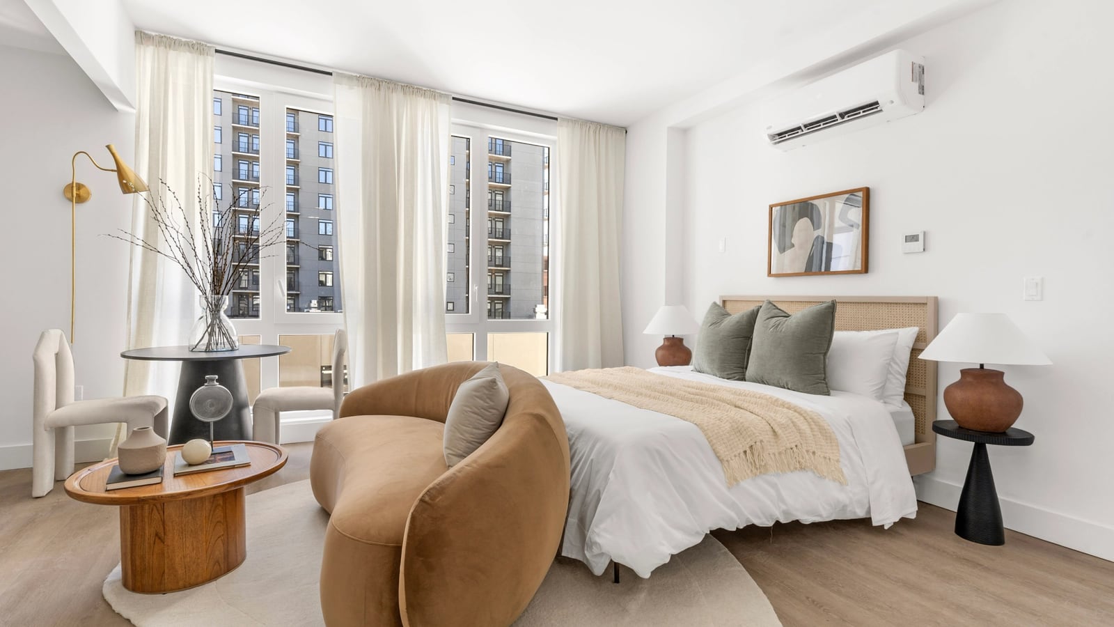 Photograph of a staged room with a bed, a curved tan sofa, a small round table and chairs, white walls and wood-look flooring, a wall of tall windows with sheer curtains looking out at a grey apartment tower with balconies, and a white wall-mounted air unit high on the wall above the bed