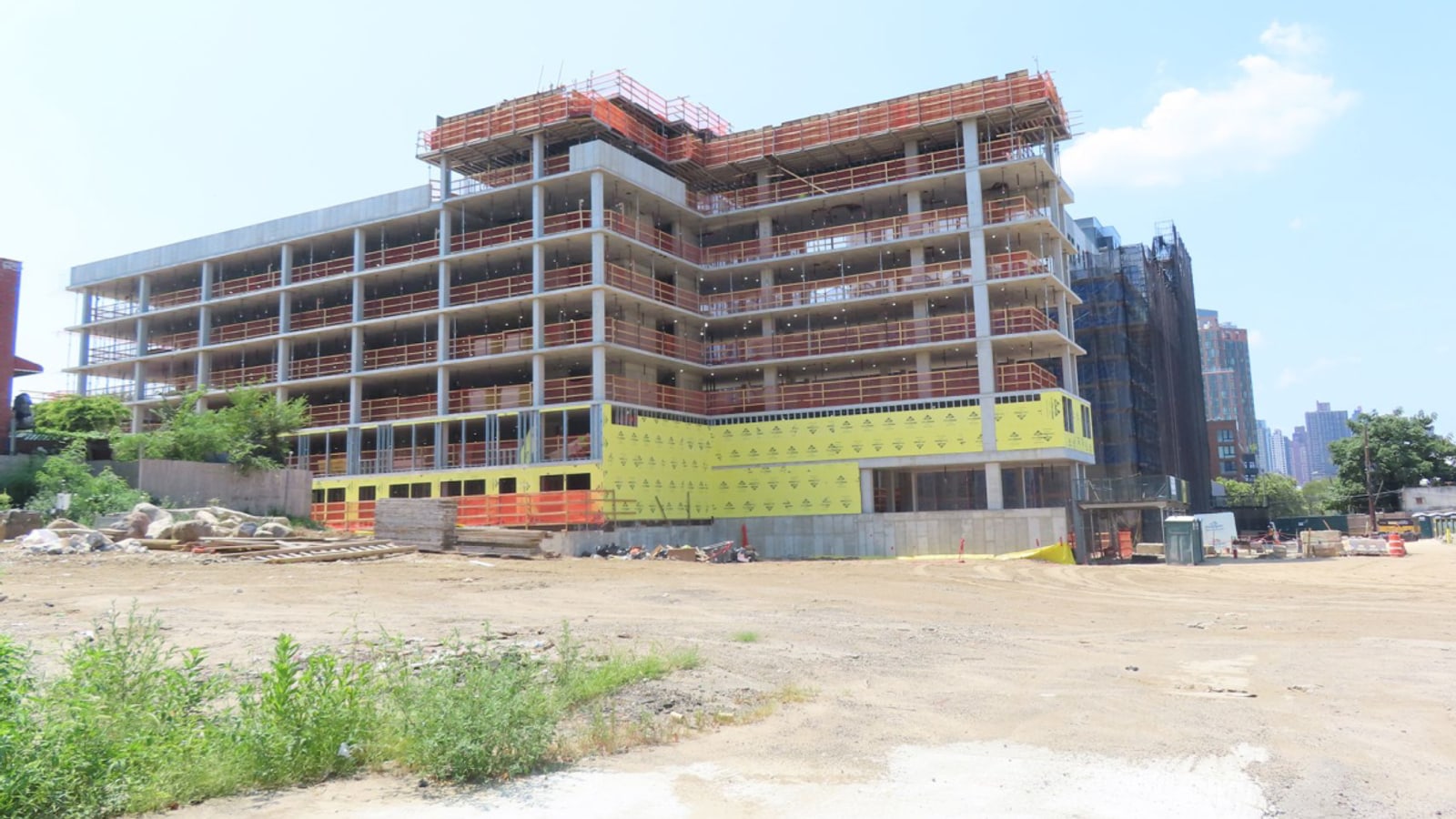Photograph of a concrete-frame apartment building under construction, with open floor slabs ringed in orange safety netting above lower floors wrapped in yellow sheathing, a second building covered in dark scaffold netting behind it at right, a bare dirt lot with weeds in the foreground and distant towers under a hazy sky