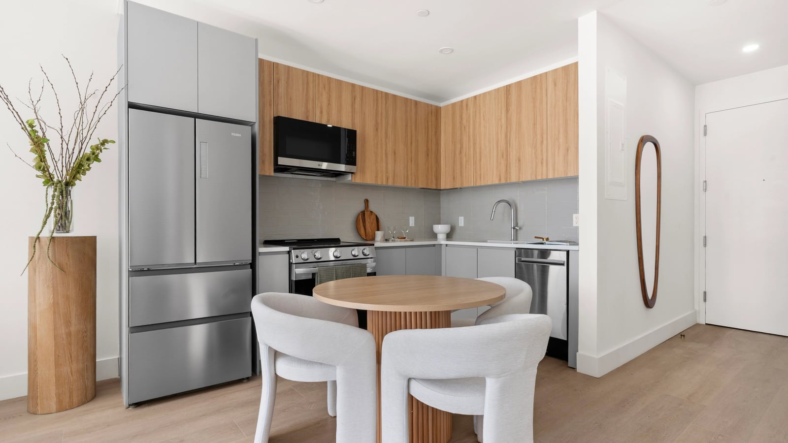 Photograph of an apartment kitchen with grey lower cabinets, wood-tone upper cabinets, a grey tile backsplash, a stainless-steel French-door refrigerator, a microwave over a range with a smooth black cooktop, a sink and a dishwasher, with a round wood table and three white upholstered chairs in front on pale wood-look flooring