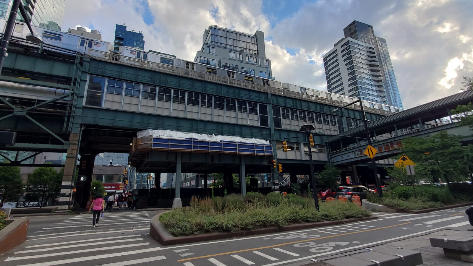 Photograph of the two-level Queensboro Plaza elevated station on green steel, with subway trains on the upper level, a planted median and bike lane in front, a crosswalk and new towers behind