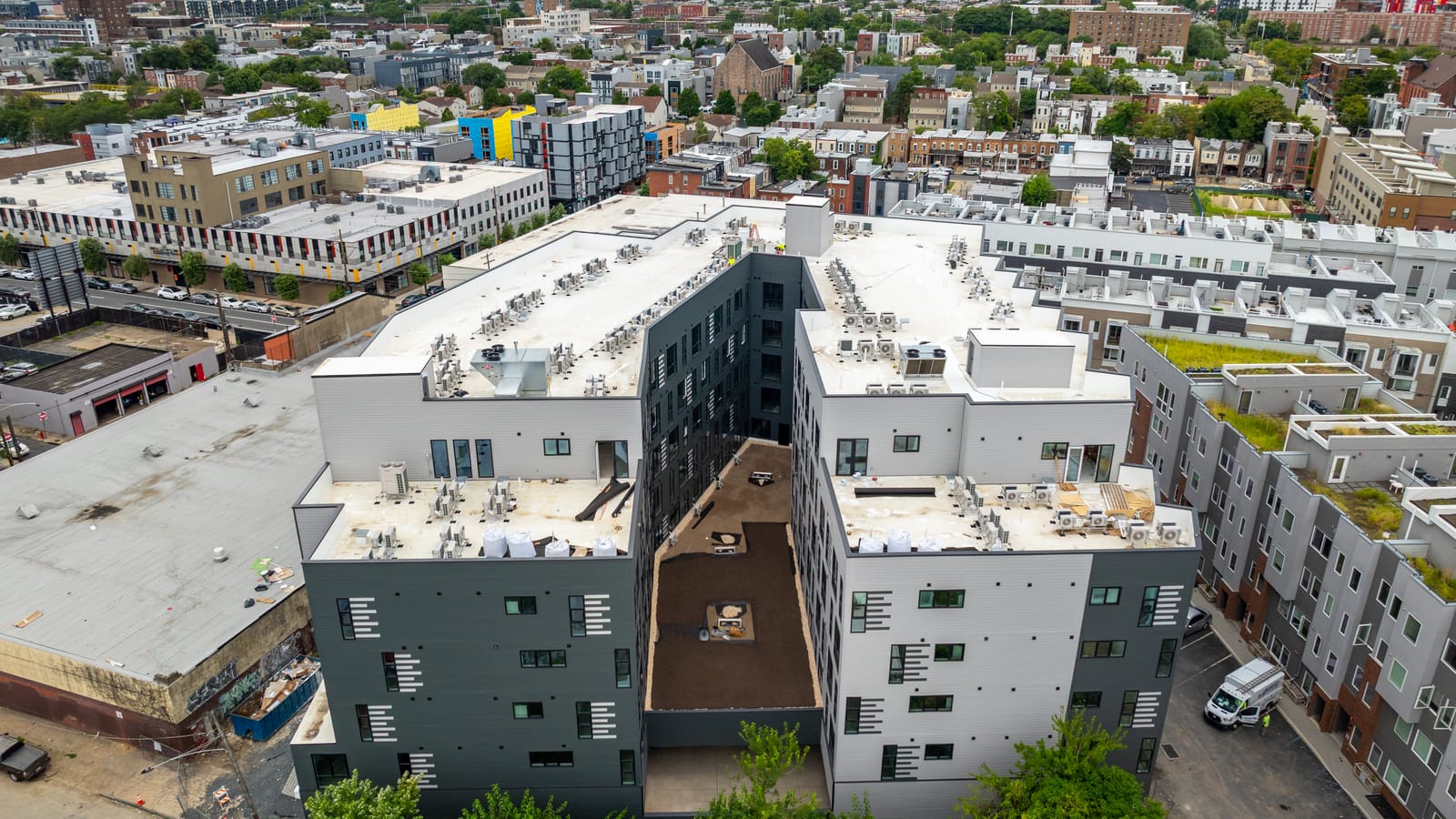 Aerial photograph of a U-shaped six-story apartment building with white roofs and rooftop equipment around a courtyard, with rows of lower buildings with planted roofs to the right and the city skyline behind