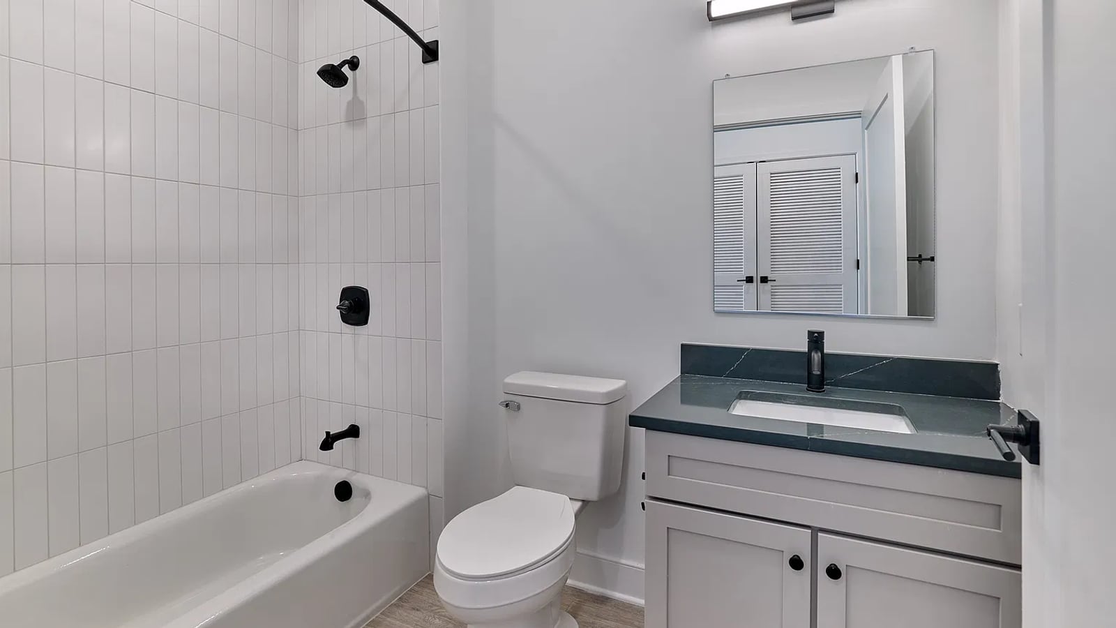 Bathroom with a bathtub and shower lined in white vertical tile, a toilet, and a gray vanity with a dark top, a black faucet and a mirror