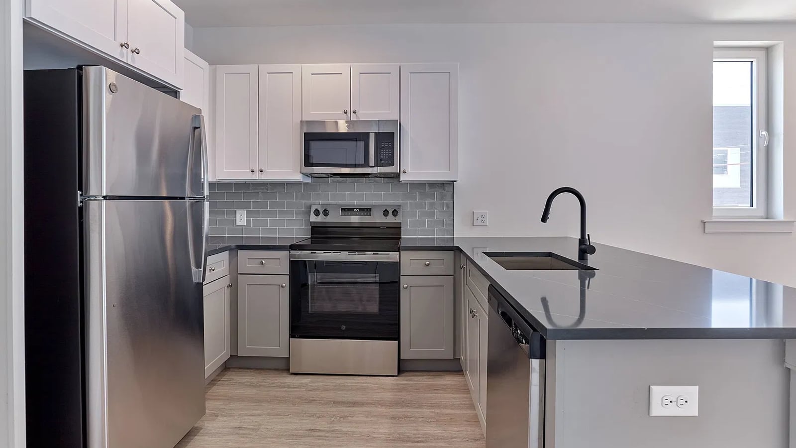 Kitchen with white upper and gray lower cabinets, a stainless steel refrigerator, electric range, microwave and dishwasher, a dark countertop with a sink on a peninsula and gray tile behind the range