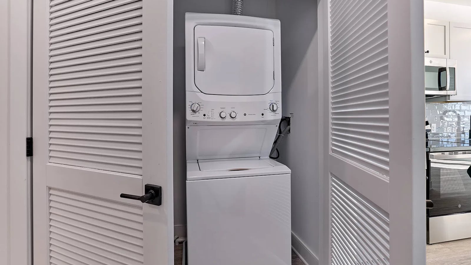 Closet with louvered double doors open on a stacked white washer and dryer, with a kitchen range and microwave visible to the right