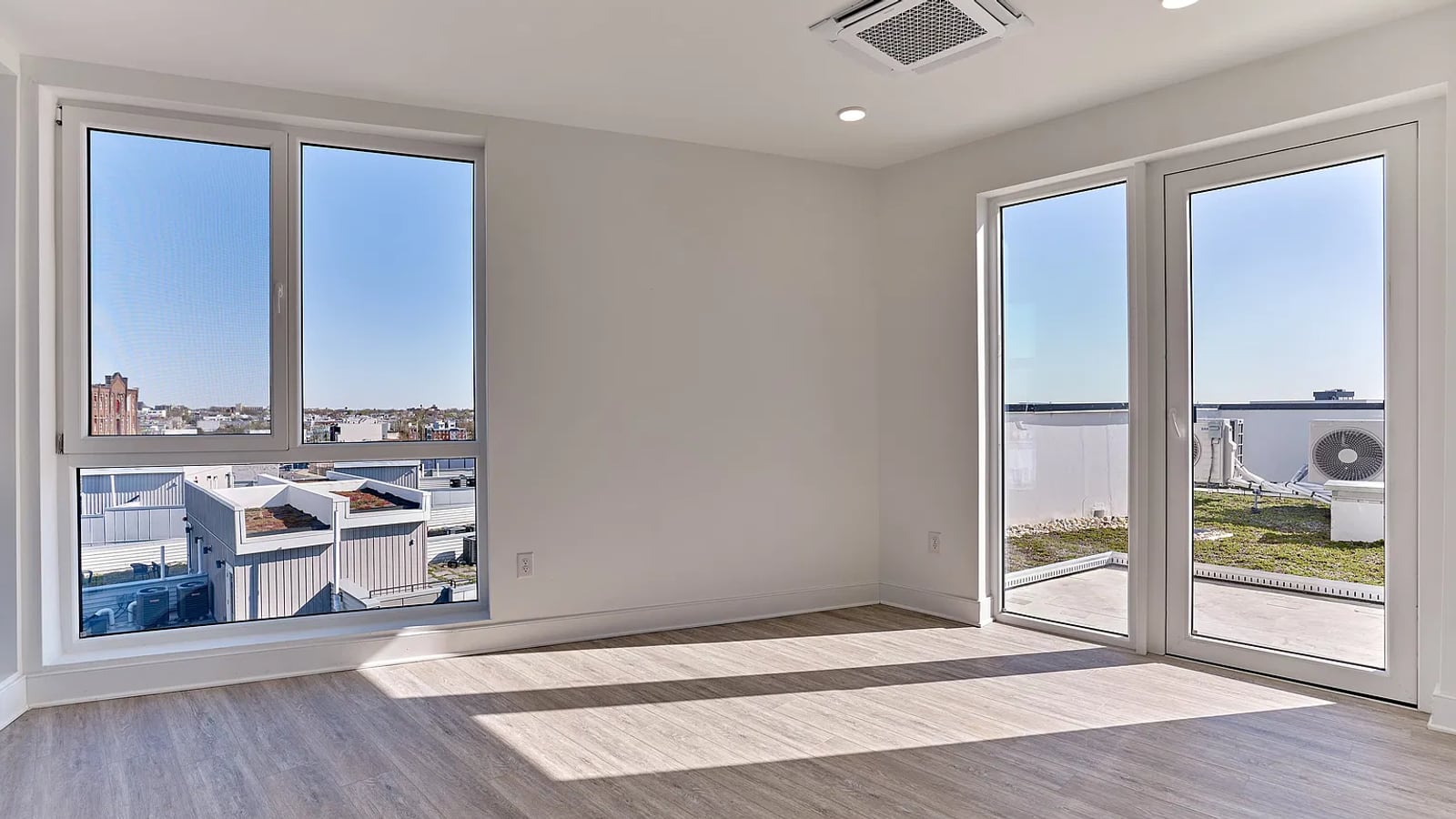 Empty room with wood-look plank flooring, white walls, a large two-part window and a glazed door opening to a terrace beside a planted roof
