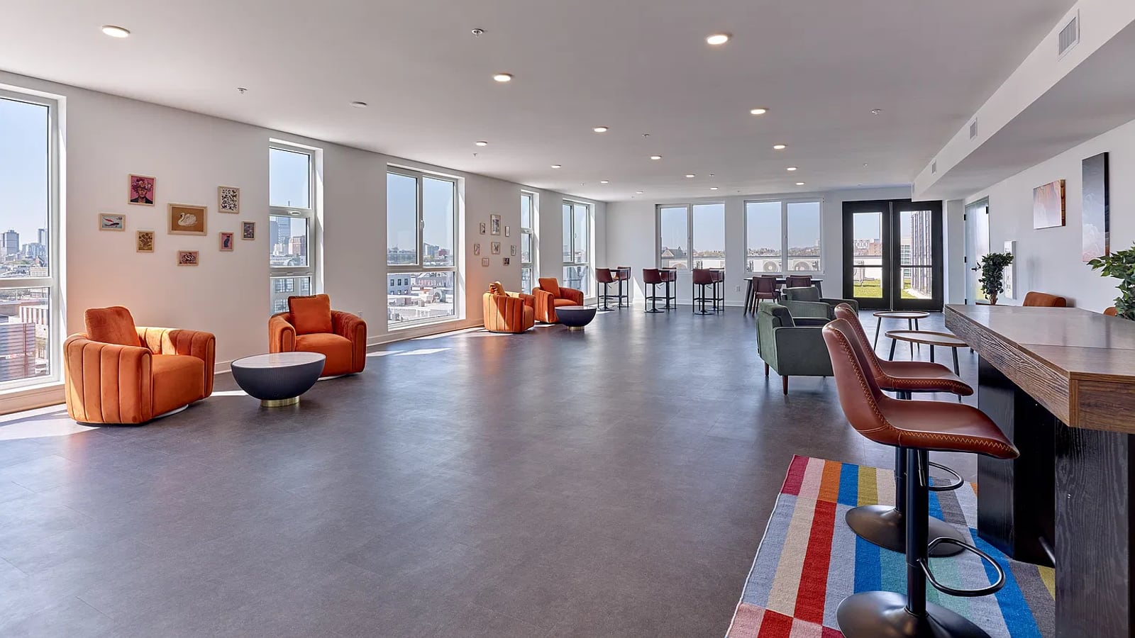 Large room with gray flooring, orange armchairs, bar stools at a long counter, a striped rug, windows on two sides and glazed doors at the far end