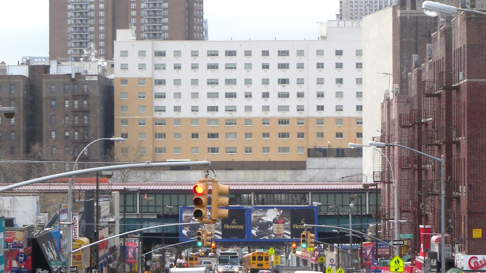 A busy street of shops and traffic lights running toward a subway station with a red roof over the street, a billboard below it and a tall white and tan building behind