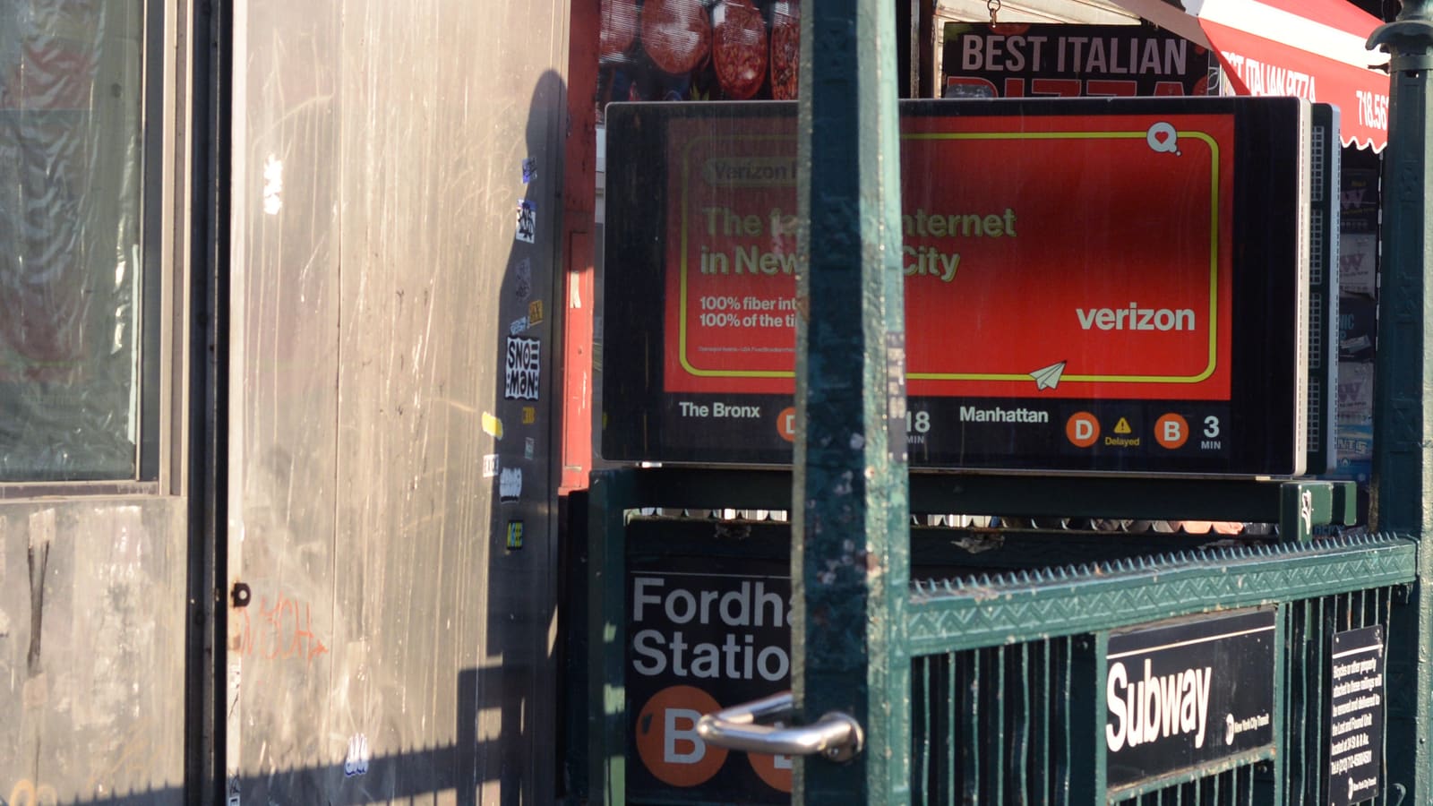 A green iron subway stair railing with a Subway sign and a Fordham Road Station sign with orange B and D circles, under a digital train-time board