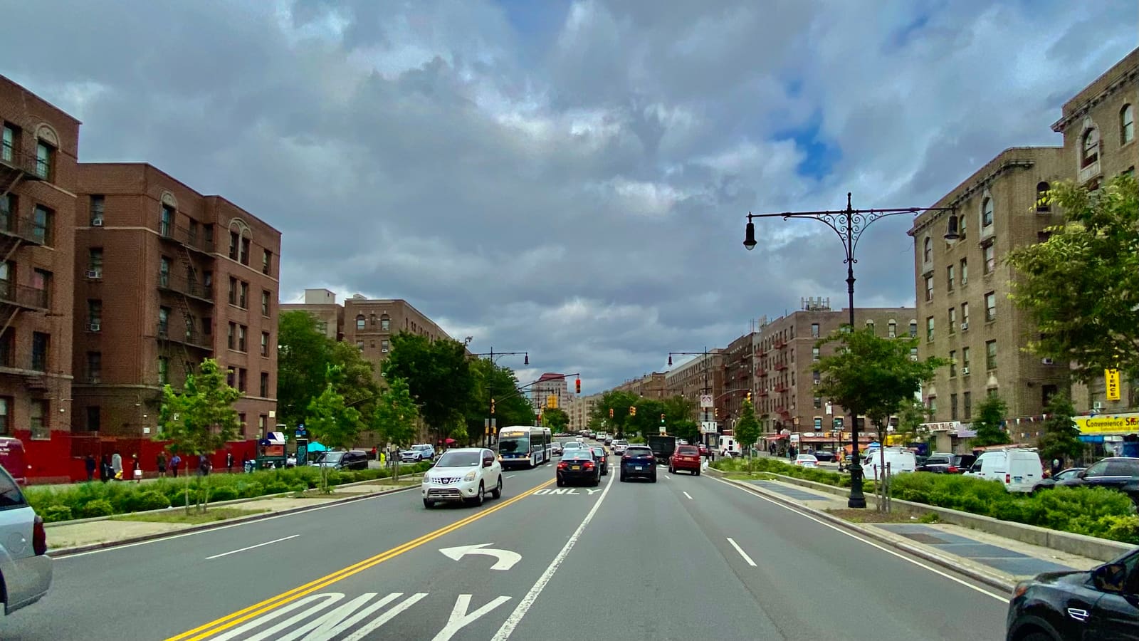 A wide avenue with a planted median, cars and a bus, lined by six-story brick apartment buildings under a cloudy sky, with ornate black lampposts