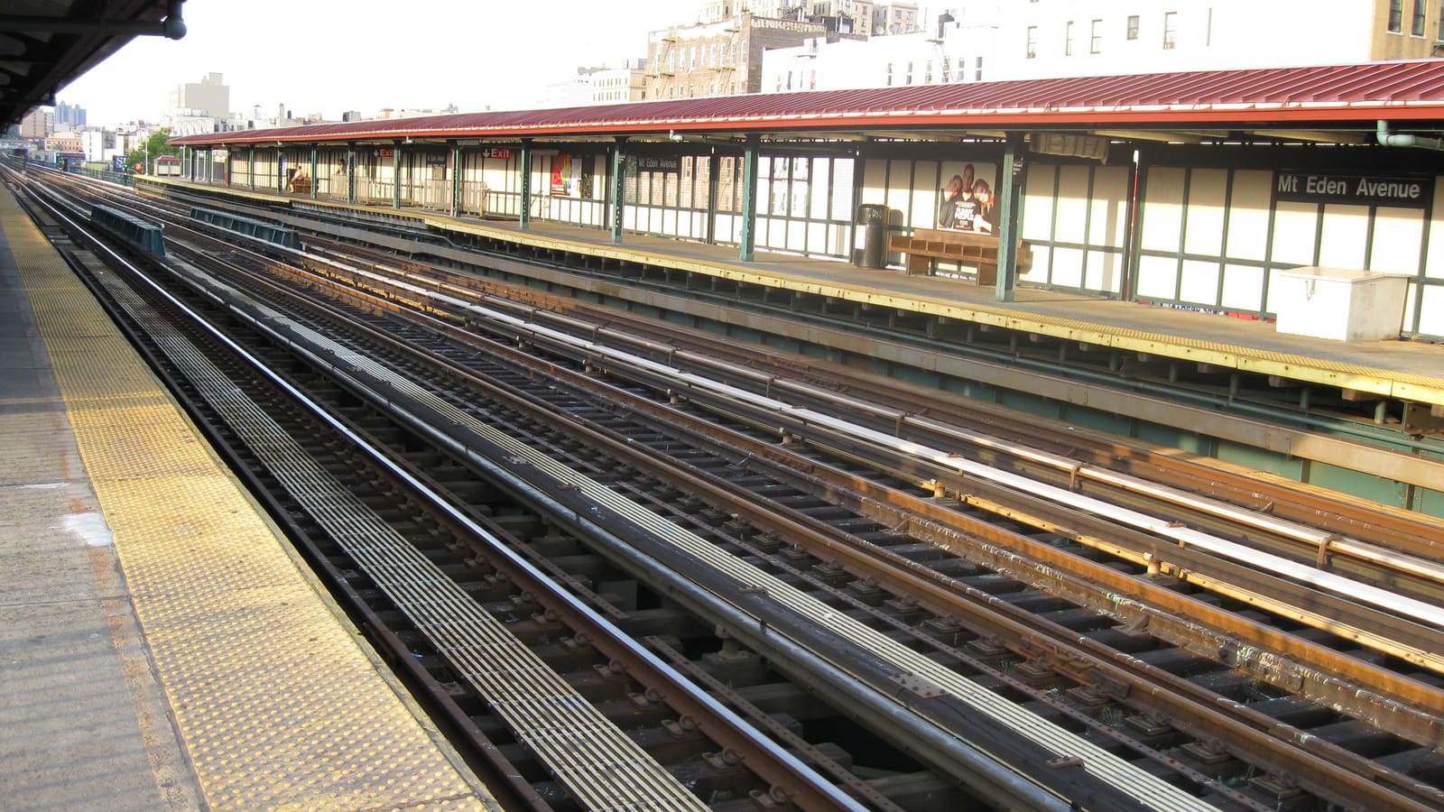 An open-air subway platform with a red roof and a Mt Eden Avenue sign, beside several sets of tracks in late afternoon light, with apartment buildings behind