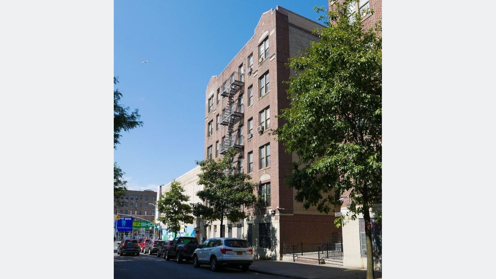 A five-story red and tan brick apartment building with a black fire escape and air conditioners in the windows, behind street trees, with cars parked along the curb under a blue sky