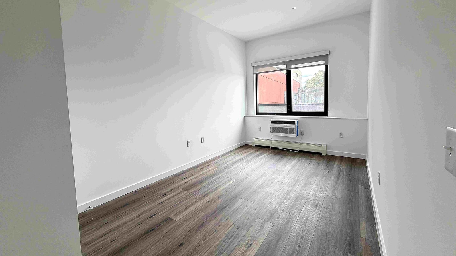 Photograph of an empty bedroom with white walls, grey-brown plank flooring, a dark-framed window with a roller shade, an air conditioner set in the wall below the window and a baseboard unit