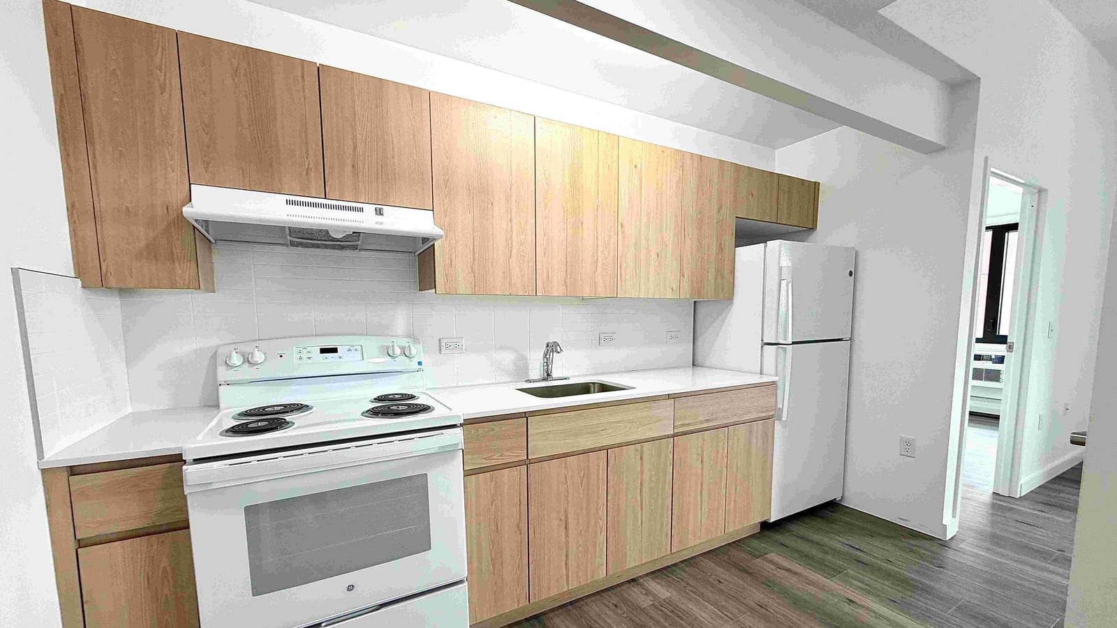 Photograph of an empty apartment kitchen along one wall: wood-tone flat-front cabinets above and below a white countertop, a white tile backsplash, a white electric range with four coil burners under a white range hood, a stainless-steel sink, a white top-freezer refrigerator and grey-brown plank flooring, with a doorway at the right to a room with a window