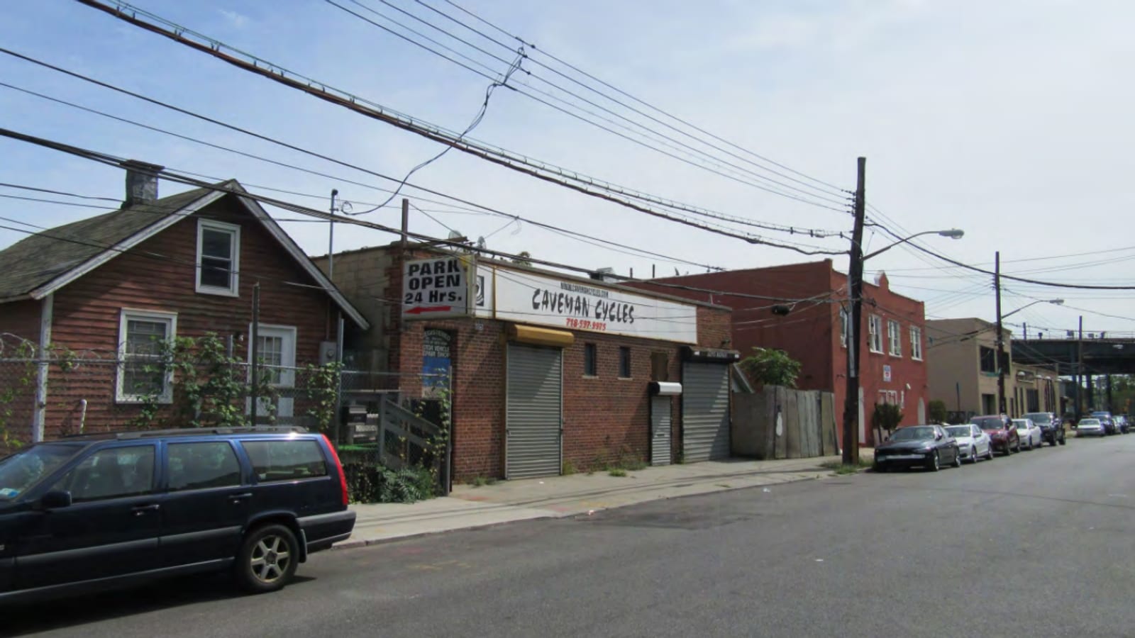 Photograph of a street with a brown wood-sided house behind a chain-link fence, a low brick building with roll-down gates under signs reading Park Open 24 Hrs and Caveman Cycles, a two-story red brick building, parked cars and overhead wires, with a raised rail structure in the distance