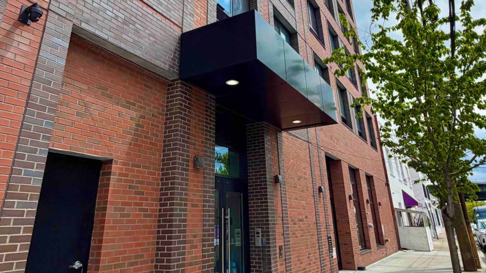 Photograph looking up from the sidewalk at a red and dark grey brick apartment building with a black metal canopy over glass entrance doors, a black service door at the left, rows of dark-framed windows above, a young street tree at the right and a low white building beyond it