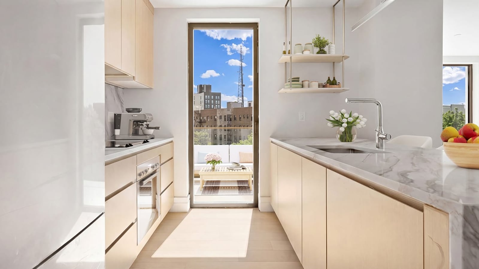 A galley kitchen with pale wood cabinets, a counter with a flush black cooktop and an oven below it on the left, an espresso machine, a marble-patterned counter with a sink and tall faucet on the right, hanging open shelves, and a glass door at the end opening onto a terrace with a view of brick apartment buildings and a radio mast