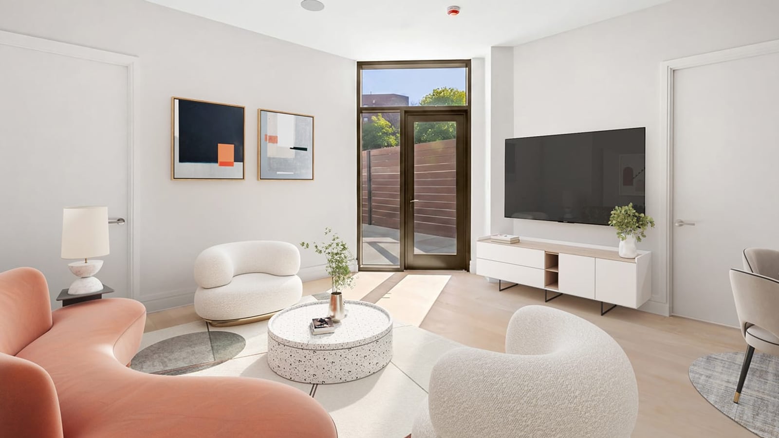 A living room with white walls, a curved coral sofa, two round white armchairs, a round speckled coffee table, a wall-mounted television above a white cabinet, two abstract prints, a dining chair at the right edge and a glass door in a dark frame opening onto a paved terrace with a wooden fence