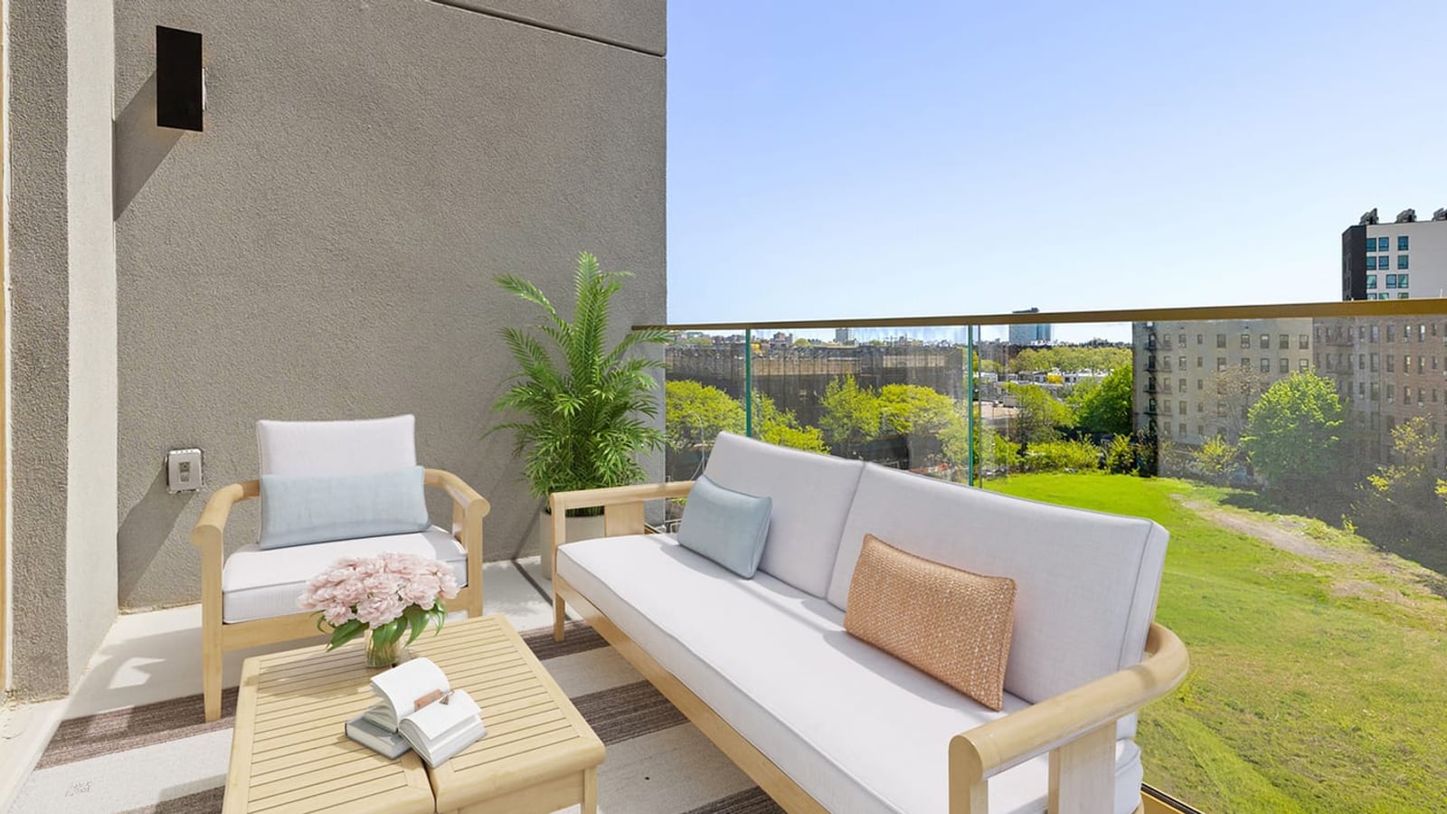 A terrace with a grey stucco wall, a wooden sofa and armchair with pale cushions, a low slatted table with flowers and an open book, a potted palm and a glass railing, overlooking a grassy lot, trees and six-story brick apartment buildings under a clear sky