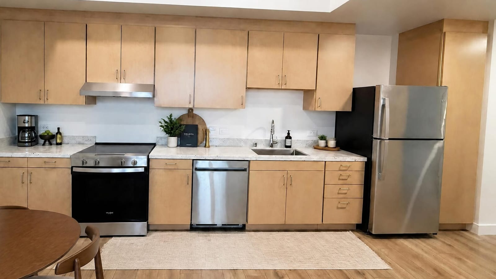A kitchen along one wall with light wood cabinets, a smooth-top electric range under a hood, a dishwasher, a sink and a stainless steel refrigerator, with part of a round dining table in the foreground