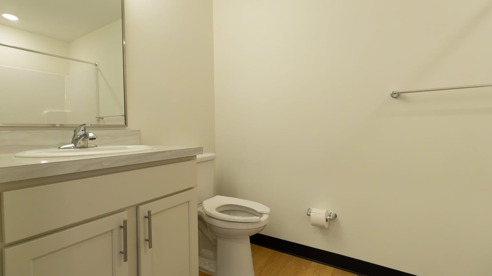 Photograph of an empty bathroom with a white vanity and sink, a mirror, a toilet, a wall-mounted bar and a wood-look floor