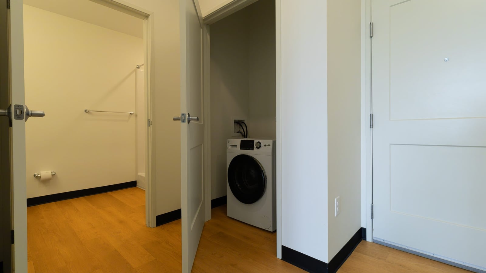 Photograph of an empty apartment hallway with a wood-look floor, an open bathroom door, an open closet holding a single white front-loading laundry machine and a white entry door