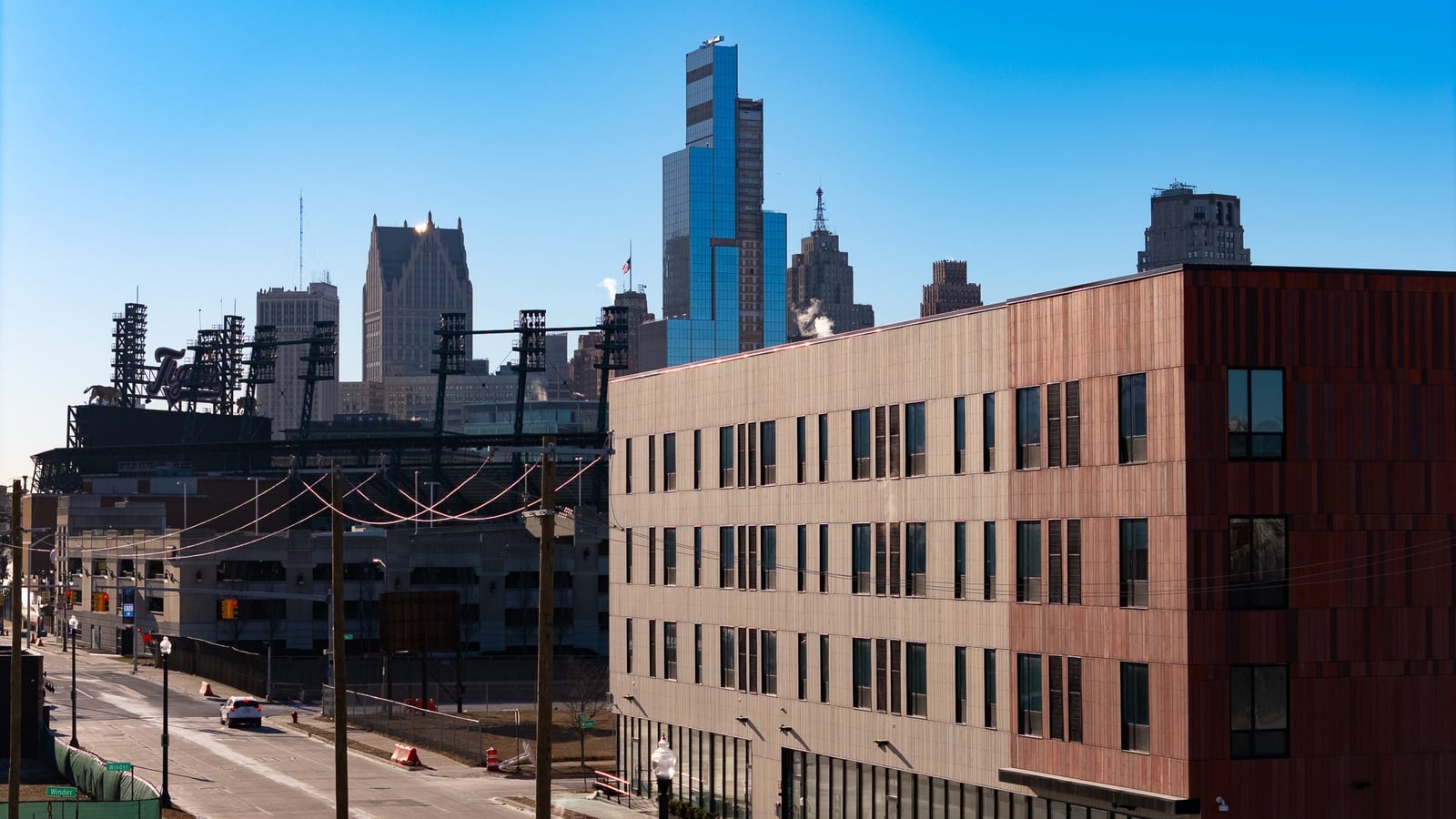 Photograph of the upper floors of a four-story apartment building with tan and red panels, with stadium light towers and downtown Detroit skyscrapers behind it
