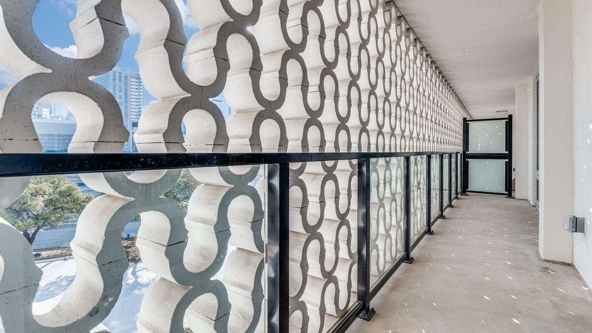 Long balcony with a glass and steel railing behind a screen of curving white concrete blocks, with towers visible through the openings