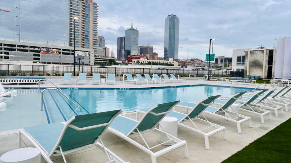 Swimming pool lined with white and teal lounge chairs, with the downtown Dallas skyline behind