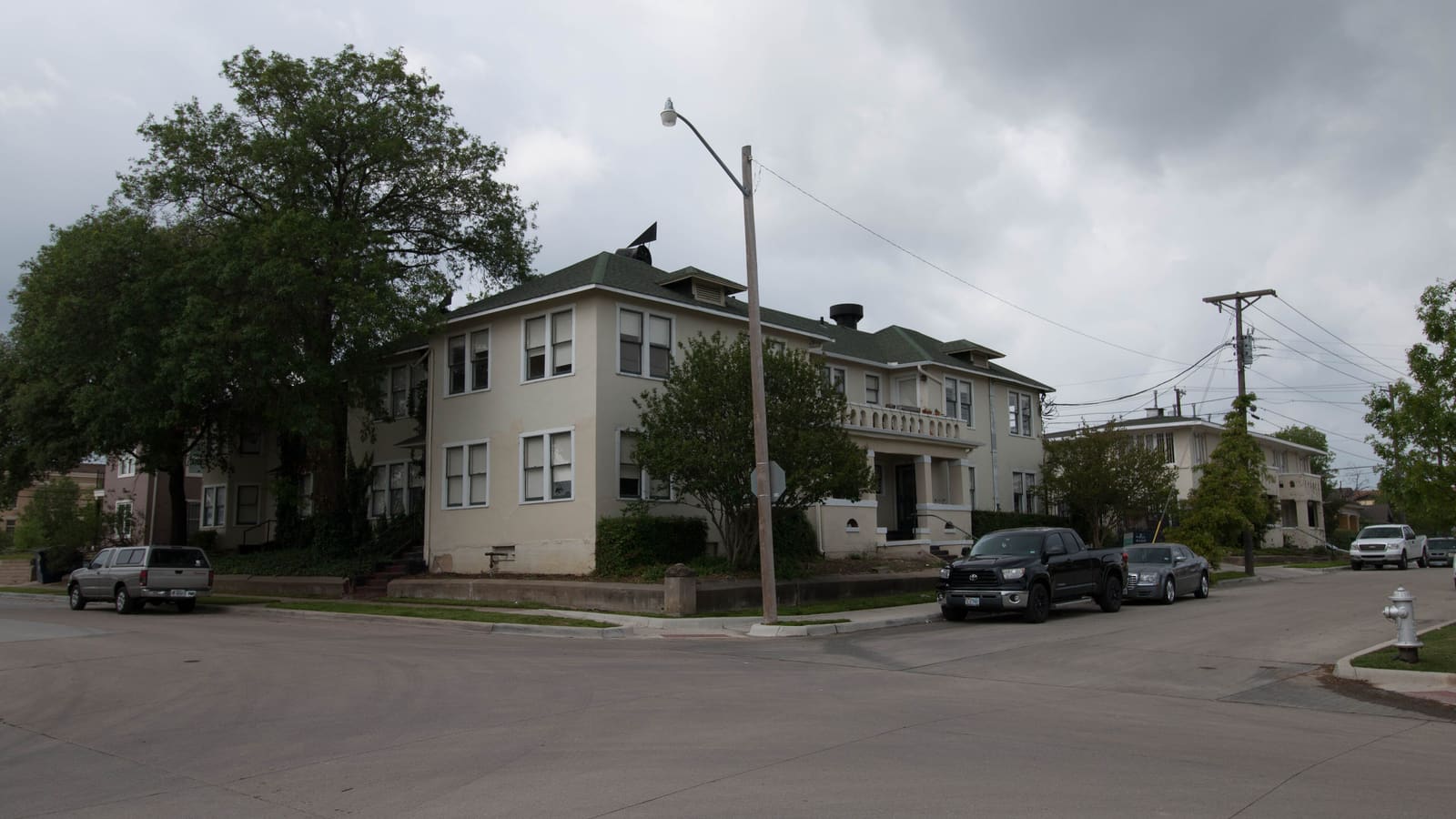 A two-storey stucco apartment building with a green hipped roof on a street corner, with trees and parked cars beside it under a cloudy sky
