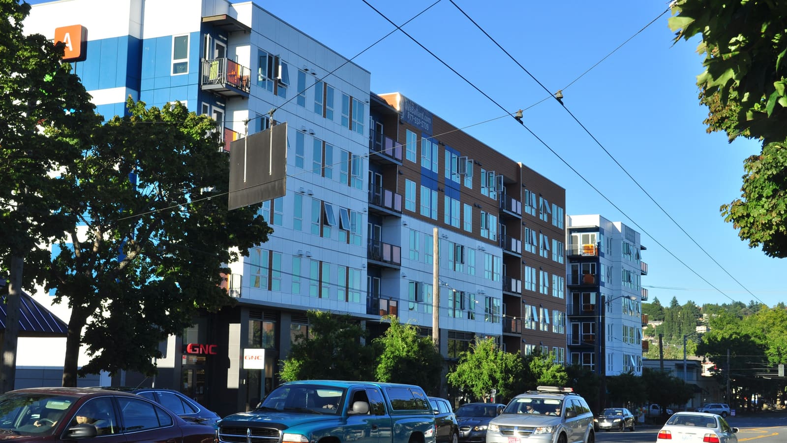 A six-storey apartment building with white, blue and brown panels and small balconies along a tree-lined street with cars passing