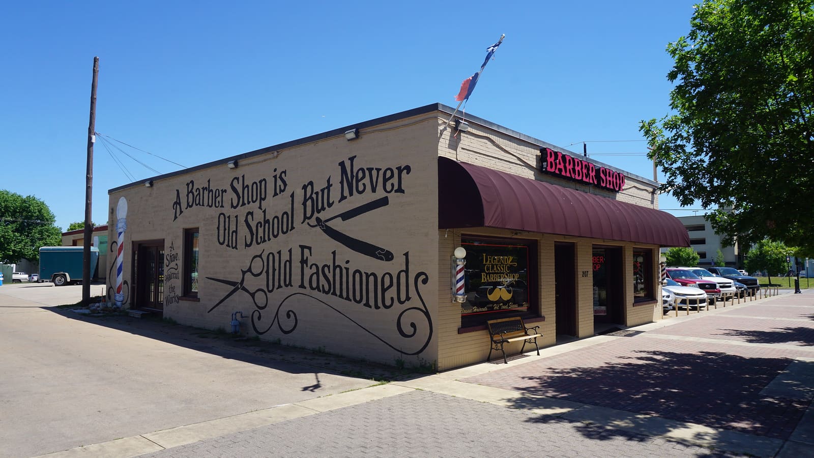 A single-storey tan brick barber shop on a corner with a maroon awning, barber poles and a painted wall sign reading A Barber Shop is Old School But Never Old Fashioned