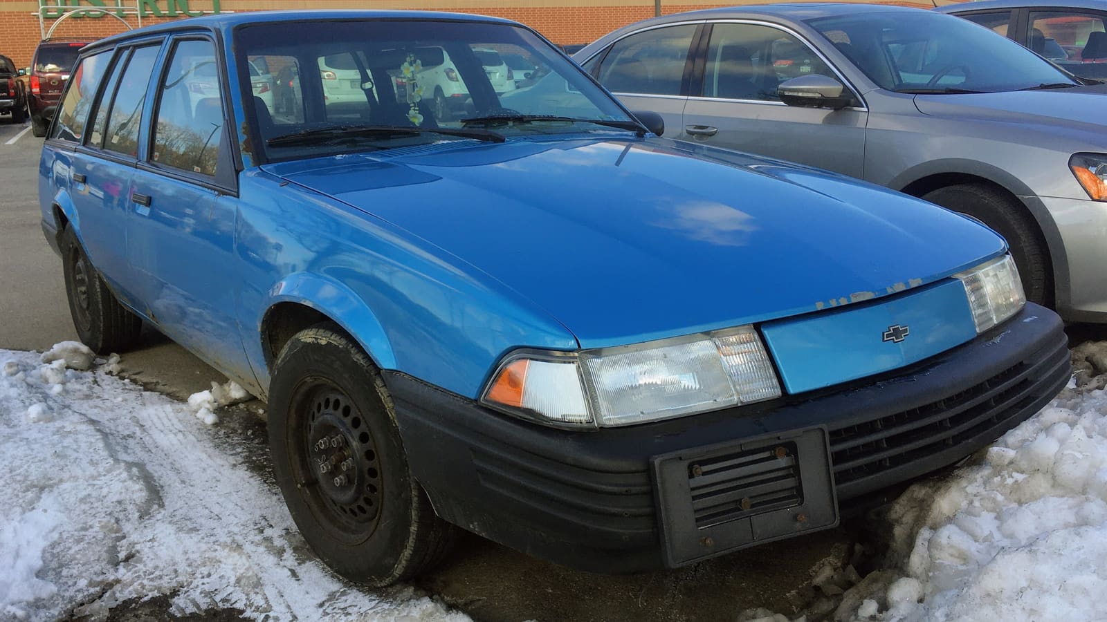 An older bright blue station wagon with plain black steel wheels parked beside a snowbank in a parking lot
