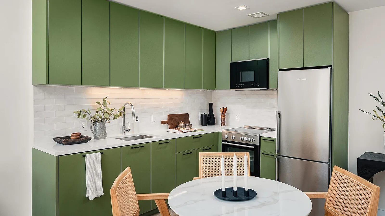 A kitchen with flat sage-green cabinets to the ceiling, a white counter and white brick-pattern backsplash, a stainless steel sink, a smooth-top electric range with front knobs, a black microwave above and a tall stainless refrigerator, with a round marble table and cane chairs in the foreground