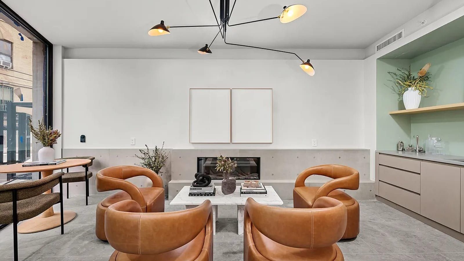 A ground-floor lounge with four curved tan leather armchairs around a white marble coffee table, a linear fireplace in a low concrete wall, a sculptural black ceiling lamp, a round wooden table with chairs by a tall street window and a mint-green kitchenette with a sink at right