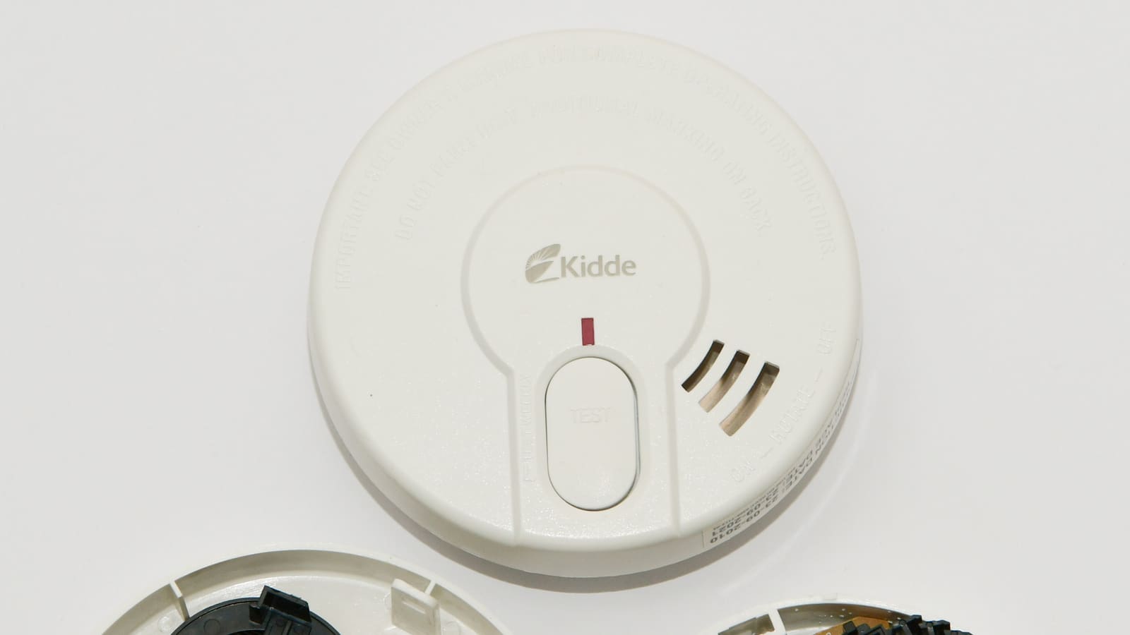 A round white household smoke alarm with a test button on its face, on a white surface
