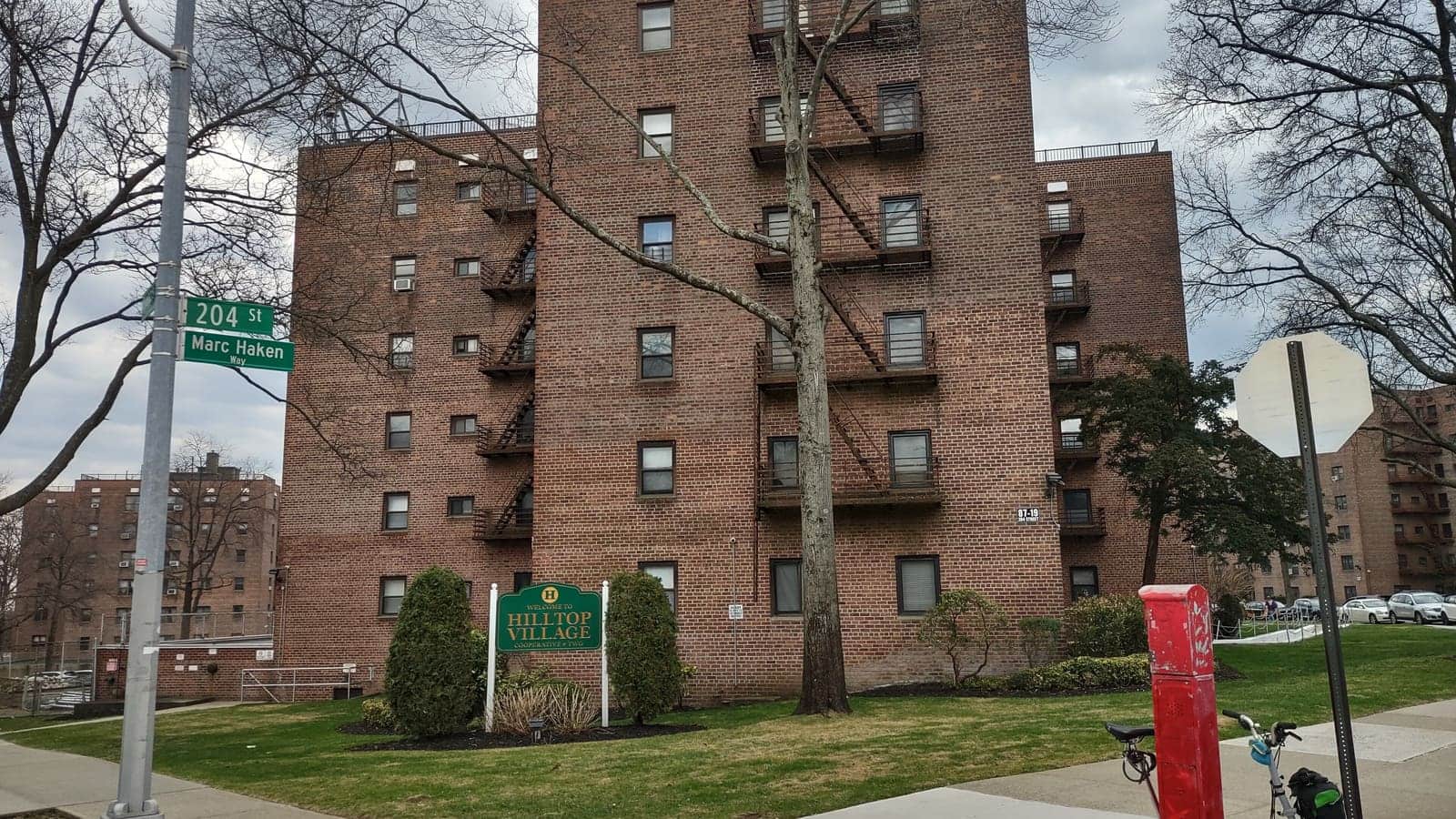 A seven-storey brick apartment building with fire escapes behind a Hilltop Village sign at the corner of 204th Street in Queens