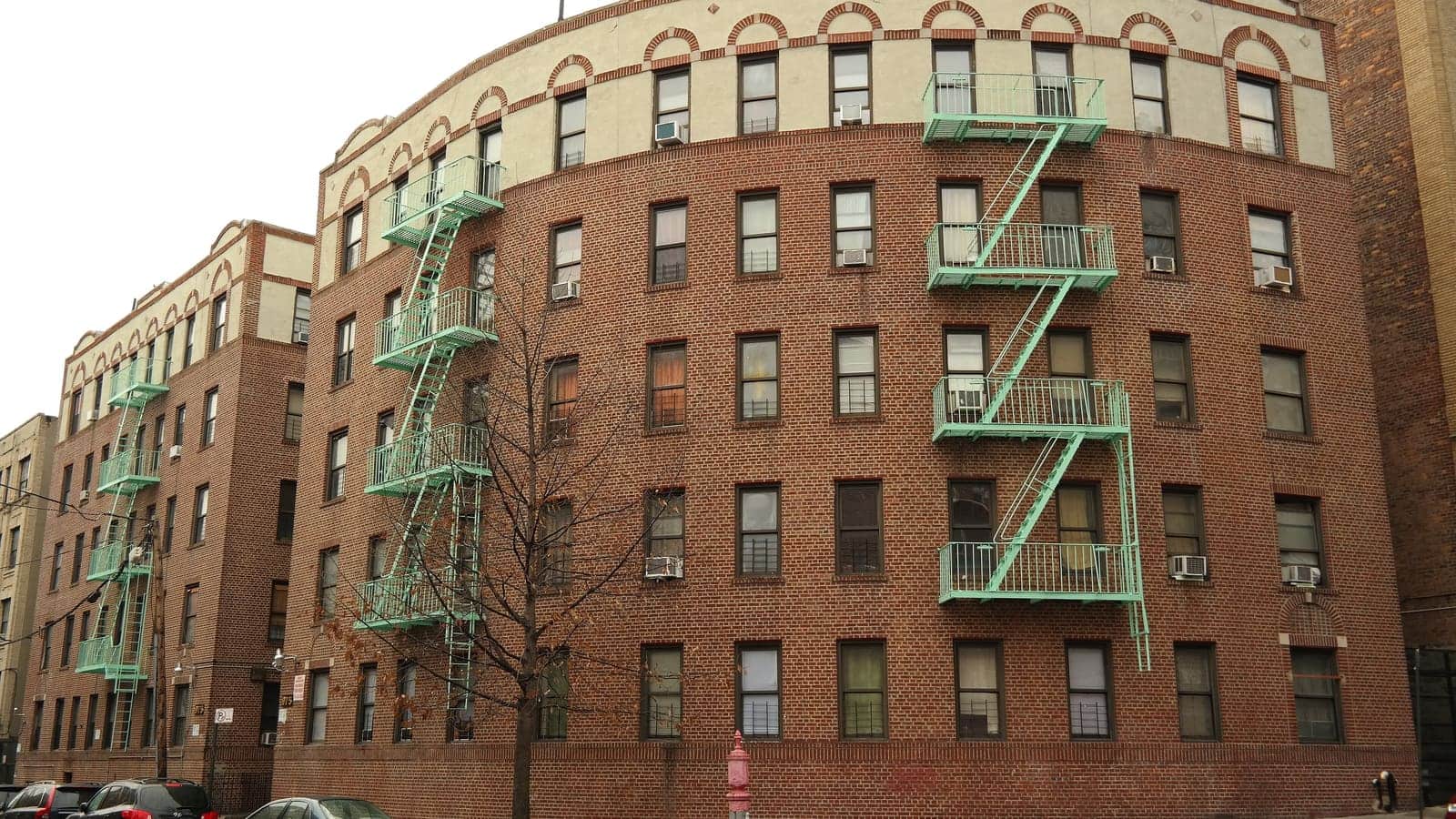A five-storey brick apartment building with green fire escapes on a curved street corner in the Bronx