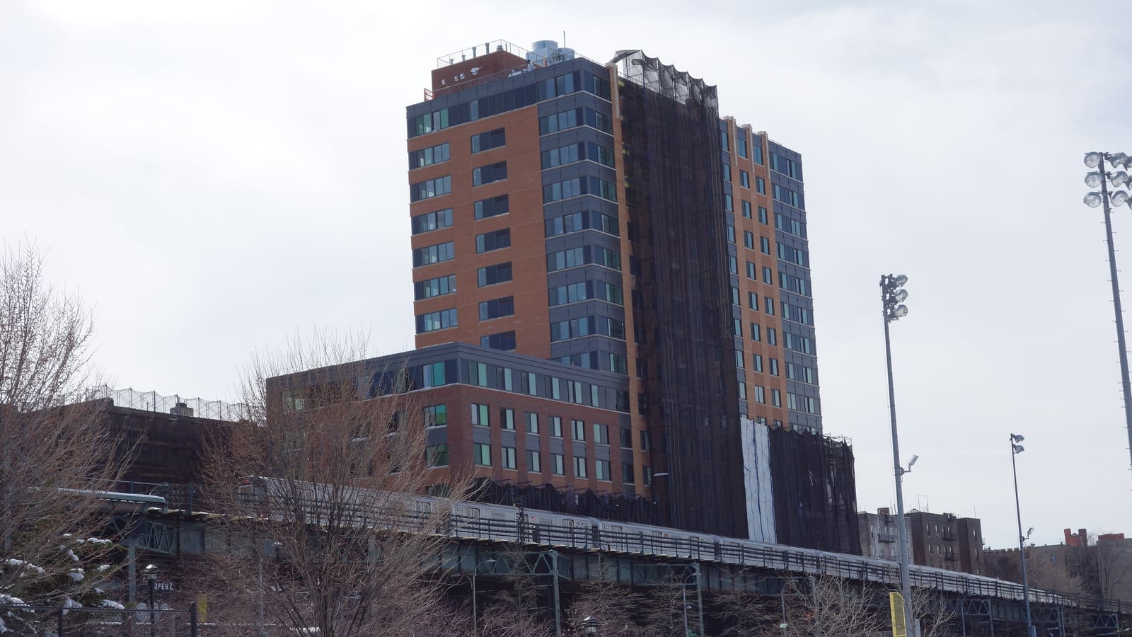 A tall brick and grey-panel apartment tower, partly covered in construction netting, rising behind an elevated subway line with a train on it, seen across a snowy park
