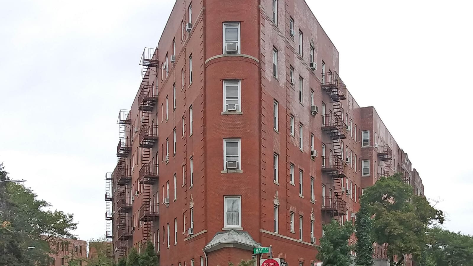 A six-story red brick apartment building with fire escapes and window air conditioners on a tree-lined street corner, with a Bay Avenue street sign in front