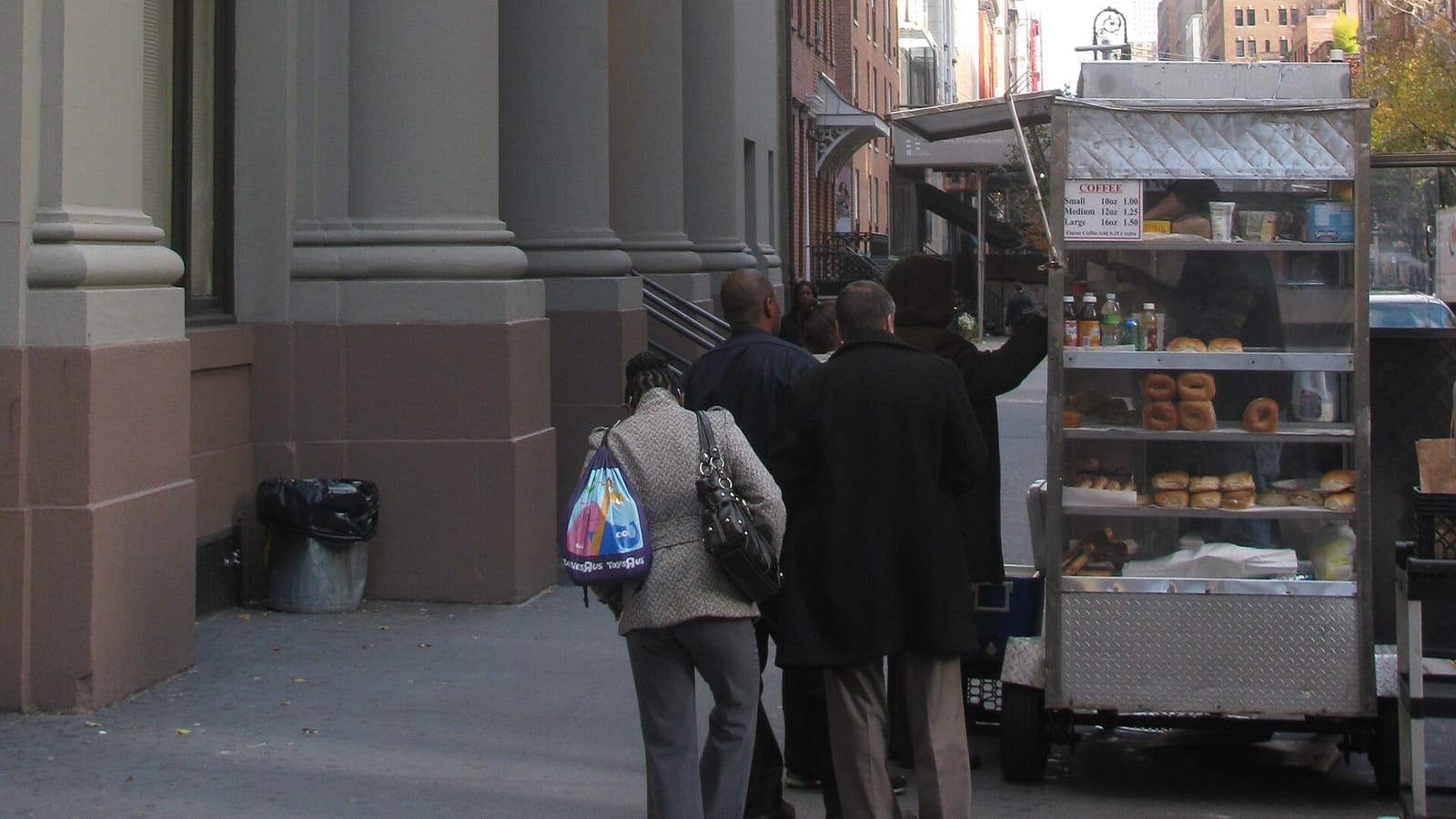 People walking past the stone columns of a government office building beside a coffee cart