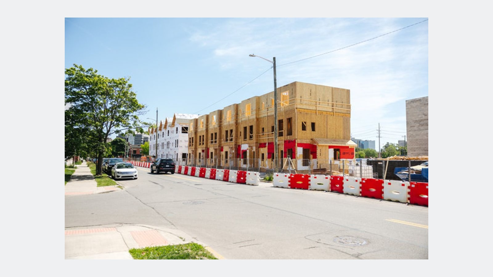 Photograph of a street with a row of three-story wood-framed buildings under construction behind red and white barriers, and a white gabled townhome at the far end