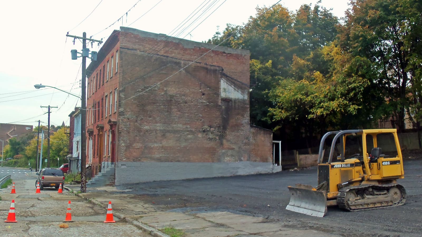 A yellow bulldozer on a freshly paved empty lot beside the bare brick side wall of a rowhouse, with traffic cones along the sidewalk