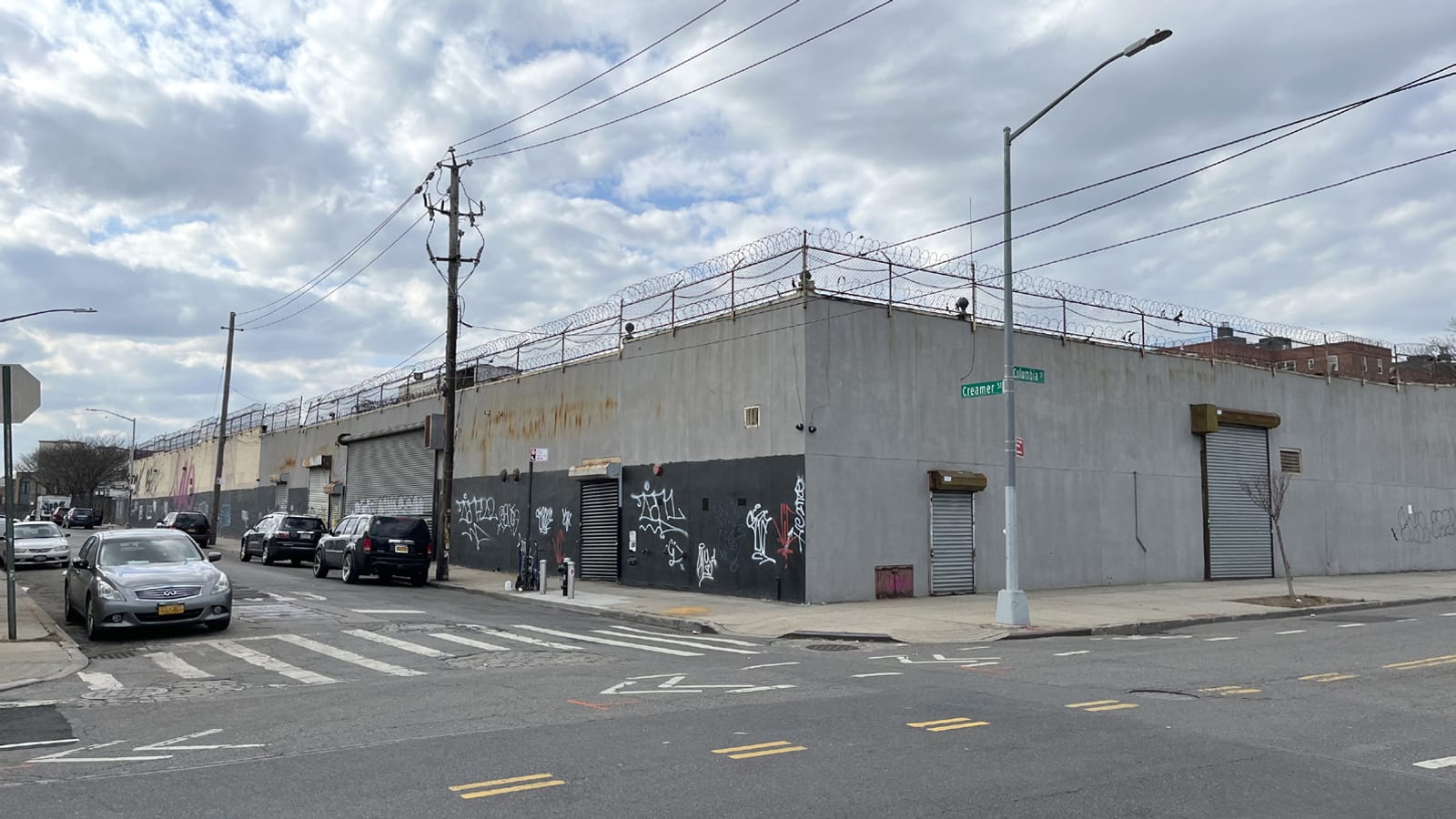 Photograph of a windowless one-story grey commercial building at a street corner, its lower wall painted black and marked with graffiti, roll-down gates shut, coiled razor wire along the roofline, a street sign reading Creamer St and Columbia St on a lamp post, and parked cars along the side street under a cloudy sky