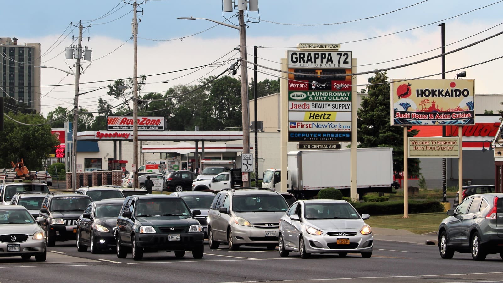 Cars waiting at an intersection on Central Avenue beside plaza signs for shops and restaurants, with utility poles overhead