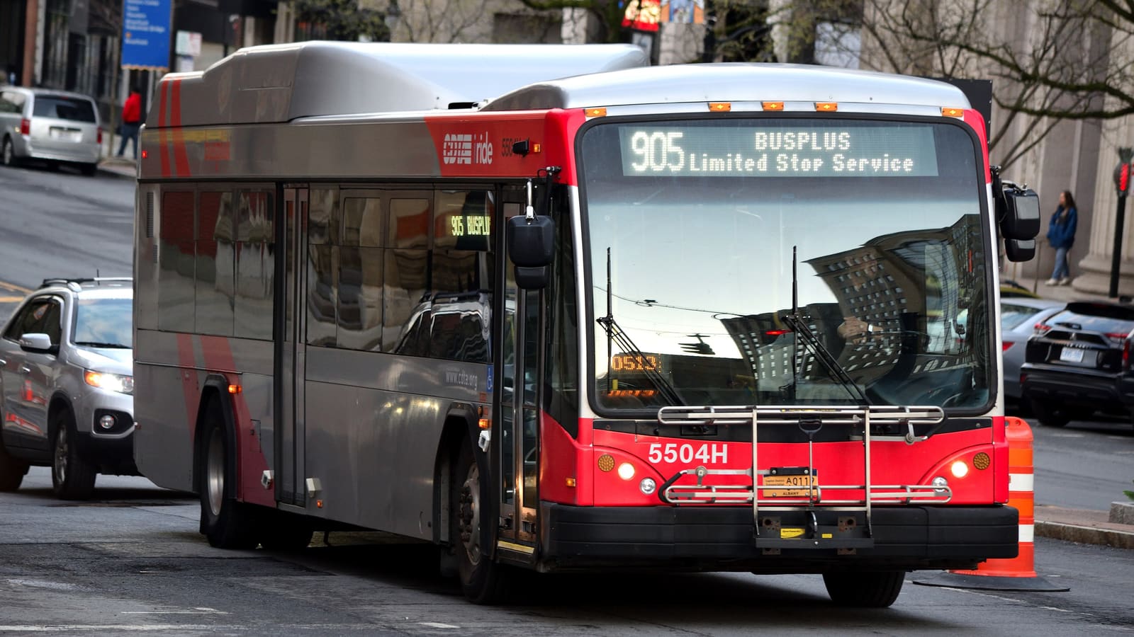 A red and silver articulated bus with a sign reading 905 BusPlus Limited Stop Service on a city street