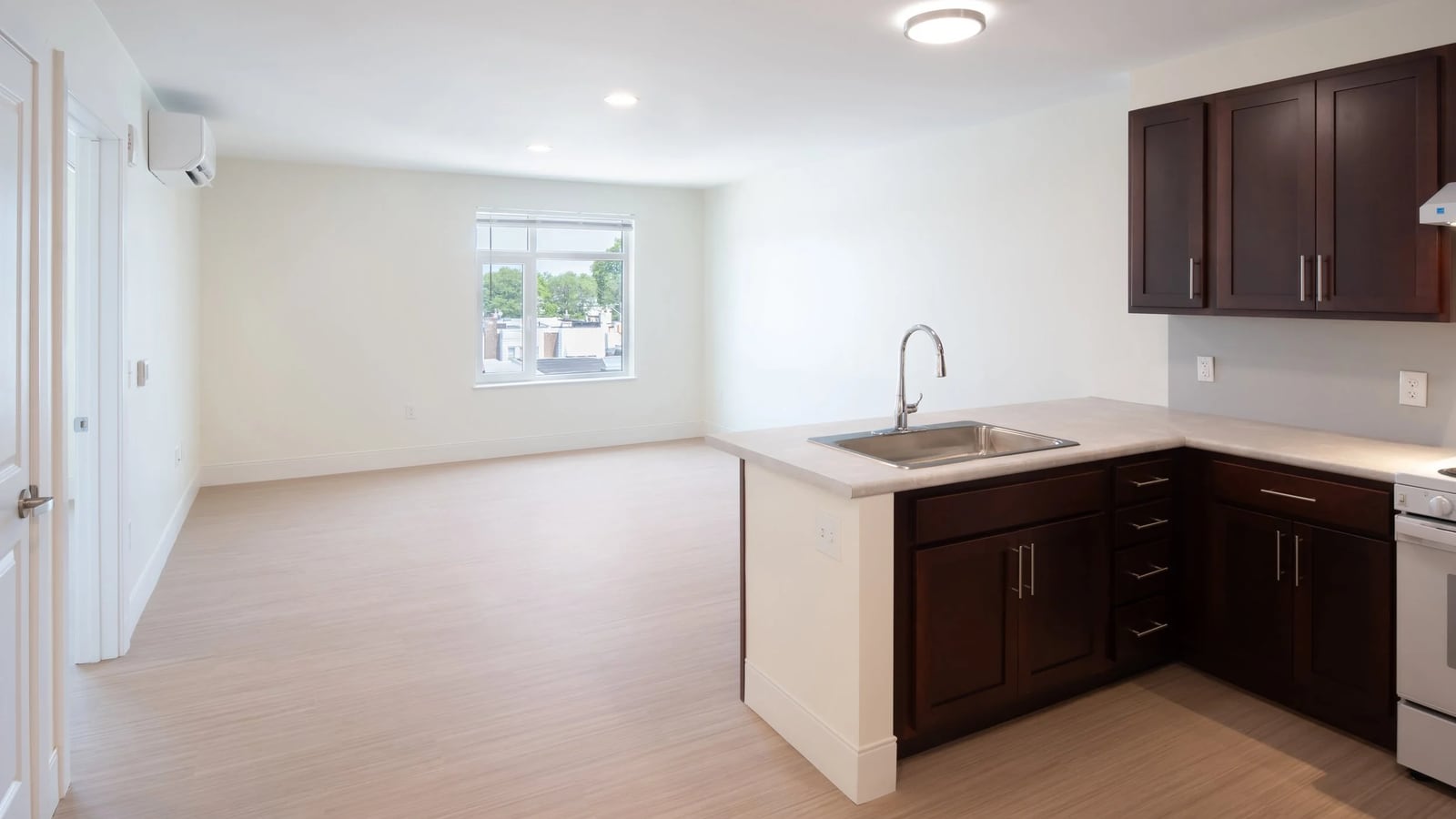 Empty apartment with light plank flooring, a window at the far wall, a wall-mounted heating and cooling unit, and a kitchen with dark cabinets and a sink set in a counter