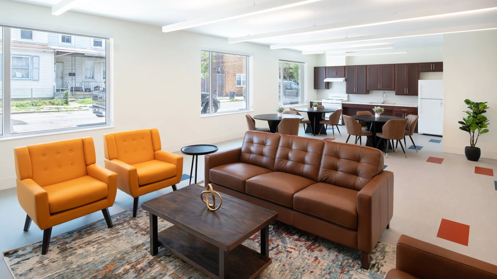 Large room with a brown leather sofa, orange armchairs, a rug, round dining tables with chairs, a kitchen with a white refrigerator and wide windows onto the street